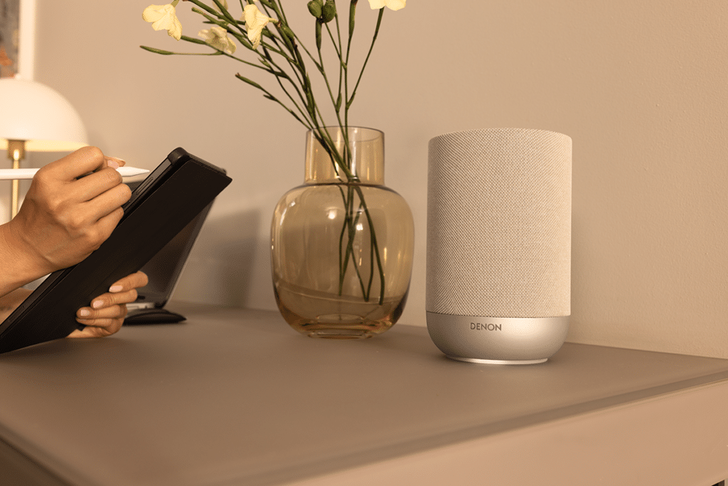 Denon Home 200 Stone -langaton HEOS/Wi-Fi-monihuonekaiutin sisustuksessa pöydällä, vaalea kangasverhoilu.